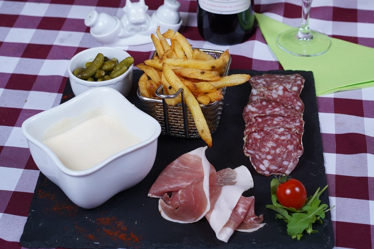 Un ramequin de fromage fondu, des frites, du jambon cru, du saucisson et des cornichons sur une ardoise